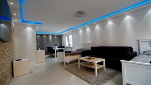 Ein modernes Wohnzimmer mit einer schwarzen Couch, einem weißen Sessel und einem kleinen Couchtisch. Es gibt eine Essecke mit Tisch und Stühlen sowie eine Küchenzeile mit Fenster.