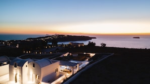 Une propriété située en bord de mer, avec un bâtiment moderne aux murs blancs, un espace repas doté de sièges en plein air, et une vue sur la mer et les collines lointaines.