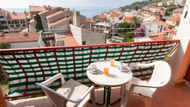 Een balkon met uitzicht op een kustplaats, met een tafel, stoelen en een gestreepte afscheiding.