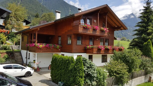 A wooden chalet with balconies adorned with flowers, surrounded by lush greenery and mountains. - Ferienhaus Pfeifer (Sankt Gallenkirch)
