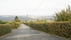 Una carretera rural bordeada de setos y árboles, que conduce a un valle con casas y una vista distante de colinas.