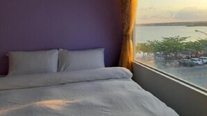 A bedroom with a bed, a window with a view of a body of water and parked cars, and a purple wall. - Hearty Homestay (Hengchun)