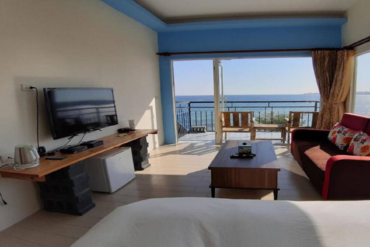 Habitació d'hotel amb llit, televisió, escriptori, cafetera, nevera petita, balcó amb vista al mar i sofà amb coixí floral.