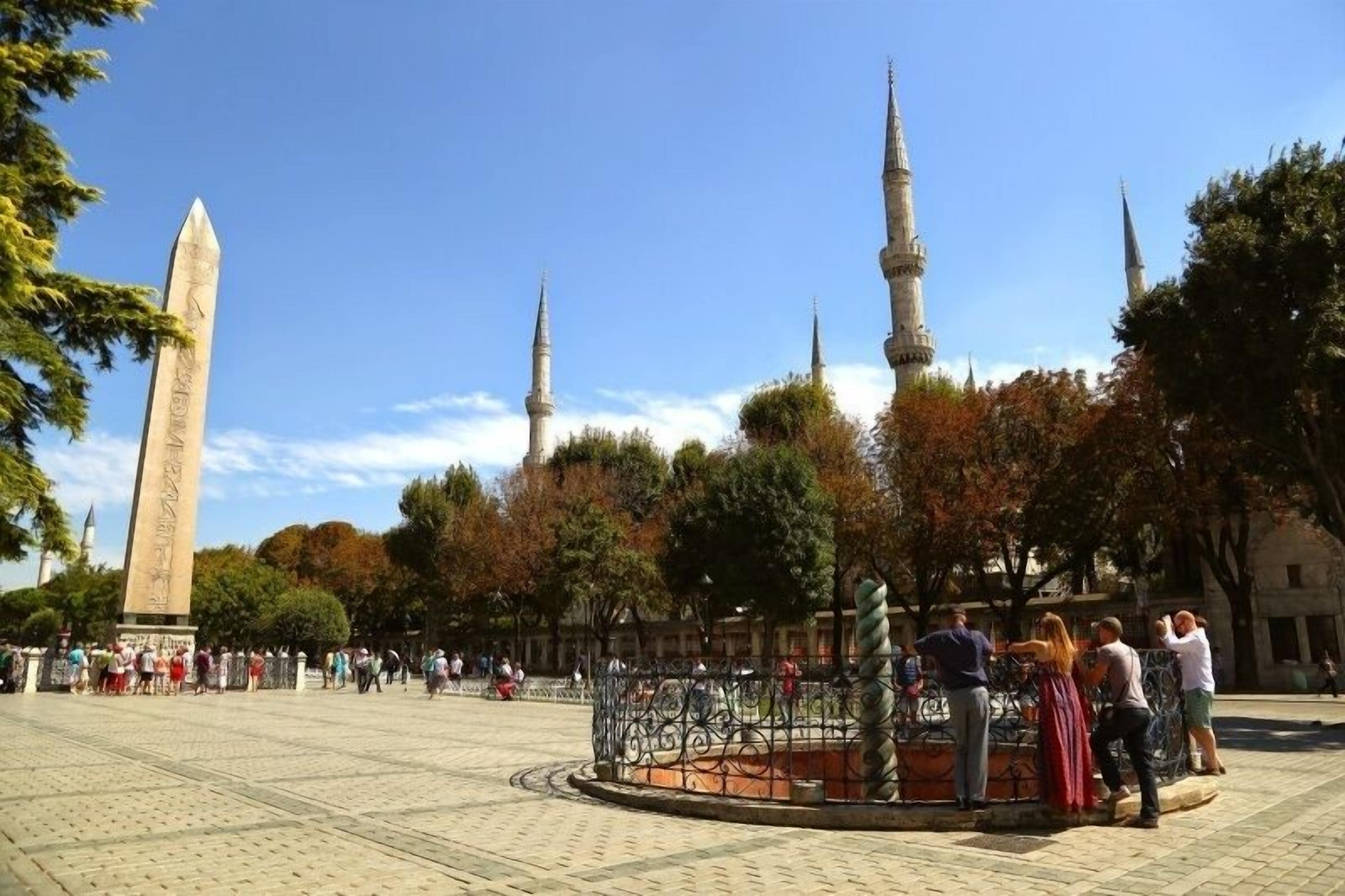 Uma praça pública com um obelisco imponente, uma fonte e minaretes ao fundo.