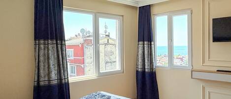 Quarto de hotel com cama, duas janelas com cortinas, televisão fixada na parede e vista para edifícios e o mar.