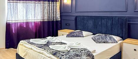 Quarto com cama, mesas de cabeceira, luz acoplada à parede e janela com cortinas.