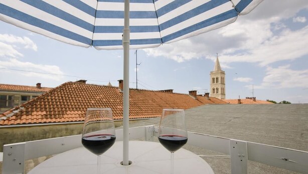 Une terrasse avec deux verres à vin placés sous un parasol à rayures, d’où l’on aperçoit un toit recouvert de tuiles et une tour.