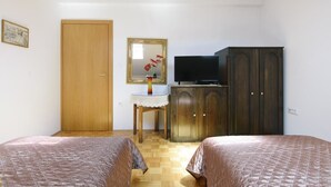 Une chambre avec deux lits, une armoire en bois, un téléviseur posé sur un meuble, une petite table avec un vase et une porte.