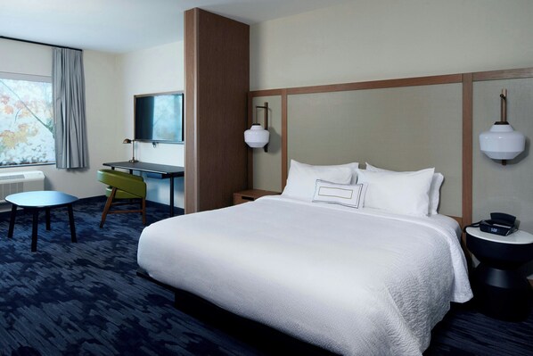 A hotel room with a large bed, a desk, a chair, a TV, and a bedside table with a lamp.