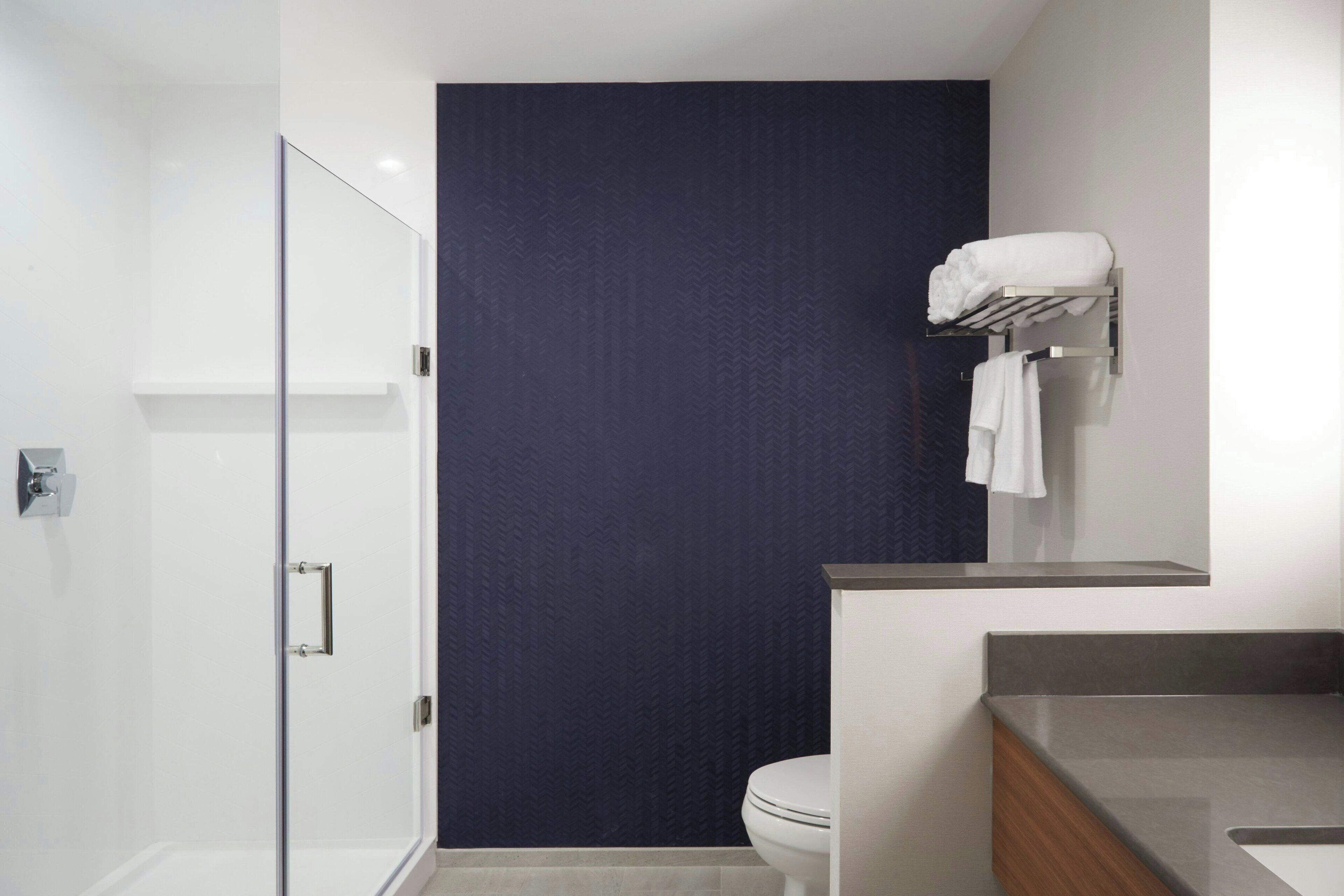 Un baño moderno con cabina de ducha de vidrio, pared de acento azul oscuro, inodoro blanco y tocador de madera con lavamanos.