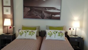 A double bed with a quilted cover, two bedside tables with lamps, a wall-mounted picture, and a small shelf with a vase. - Top Floor Cozy Apartment near Beach (Vari-Voula-Vouliagmeni)