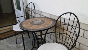 Een klein terras buiten, met een ronde tafel en twee stoelen, een deur met deurmat en een betonnen vloer.
