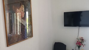 Een kamer met een groot schilderij in lijst, een flatscreen televisie aan de muur, een zwarte speaker en een klein tafeltje met plantenpotten.