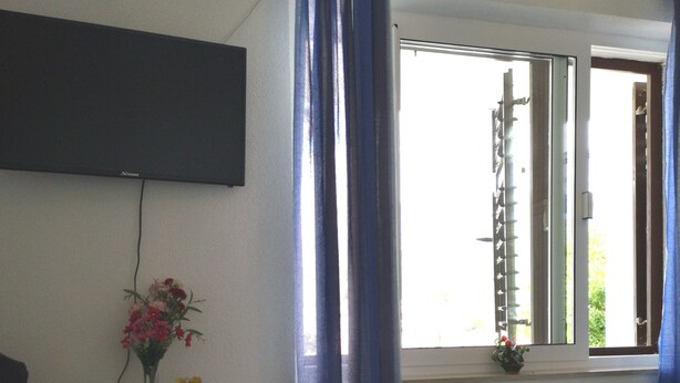 Een slaapkamer met een bed, een dressoir met een flatscreen televisie erboven, een raam met gordijnen en een kleine plant in een pot op de vensterbank.