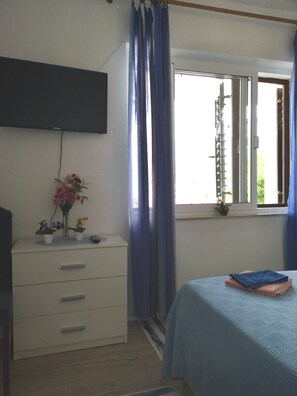 A bedroom with a bed, a dresser with a flat-screen TV mounted above it, a window with curtains, and a small potted plant on the windowsill.