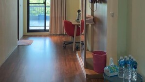 Une pièce spacieuse avec un plancher en bois, une porte-fenêtre coulissante donnant sur un balcon, une chaise rouge, une petite table avec des bouteilles d’eau et une poubelle.