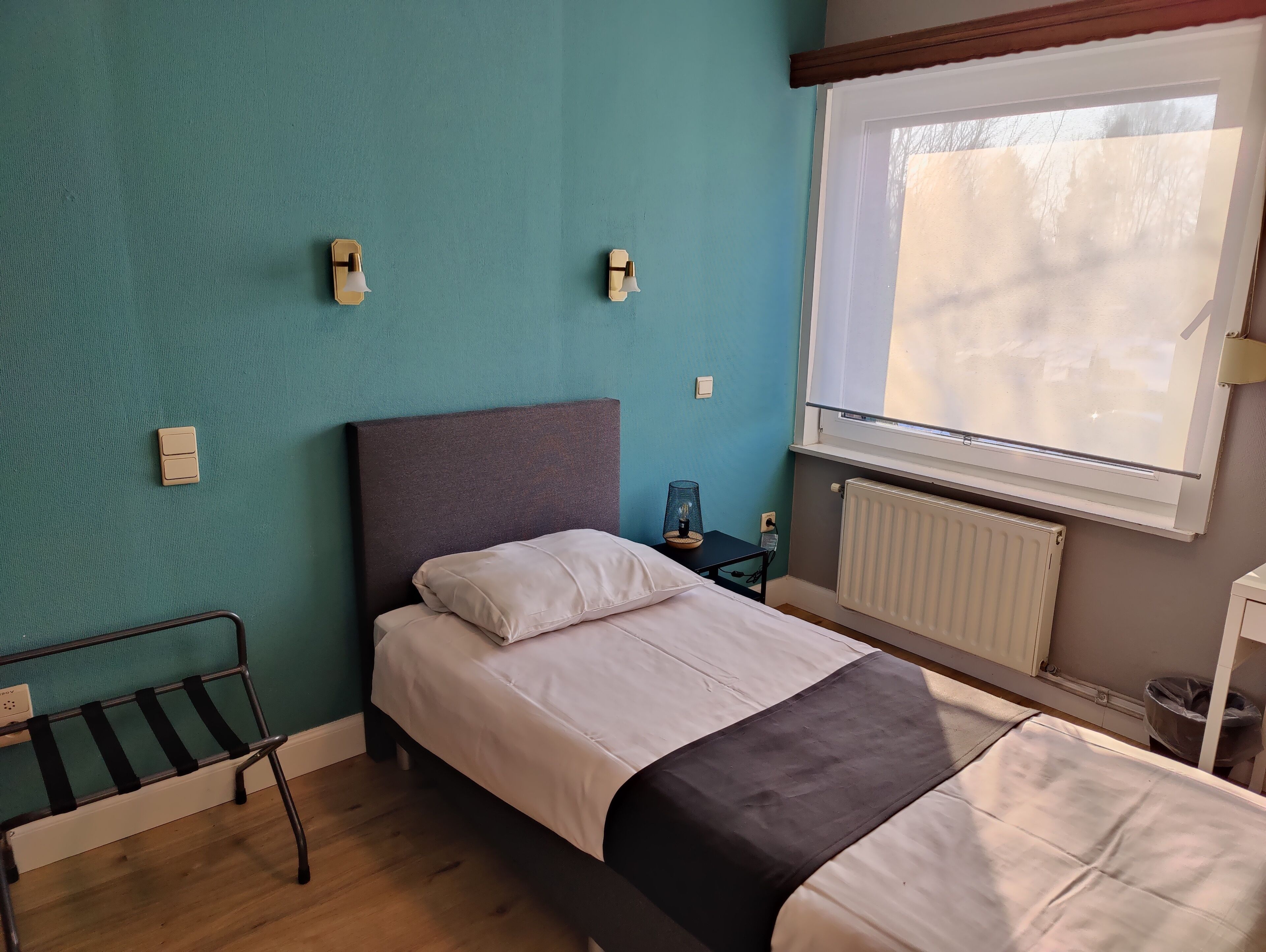 Una habitación con una cama, un banco, una mesa con lámpara y una ventana con persianas.