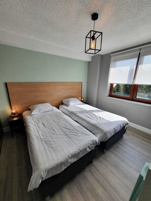 Un dormitorio con dos camas, cabeceros de madera, una mesita de noche con lámpara y una ventana con persianas.