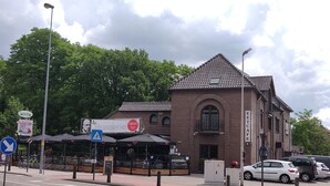 Een gebouw met een pannendak en rondboogvensters, naast een straat met geparkeerde auto's en een café met terras.