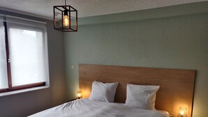 Een netjes opgemaakt bed in een kamer met een houten hoofdeinde, twee bedlampen en een raam met jaloezieën.