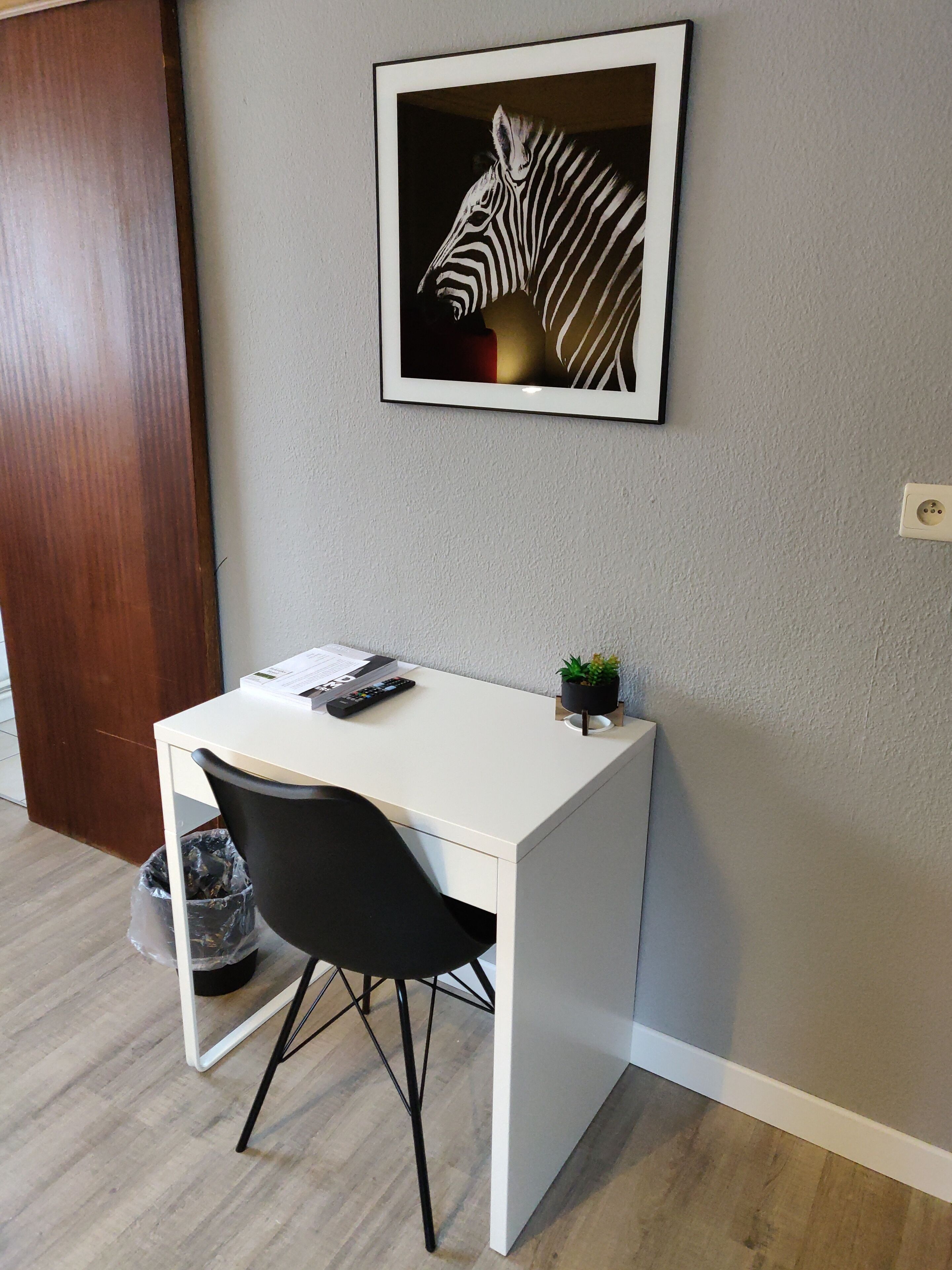 A small, white desk with a black chair, a framed zebra artwork on the wall, and a small plant on the desk.