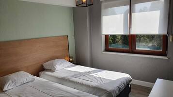 Een slaapkamer met twee bedden, een houten hoofdbord, een nachtkastje met een lamp, een raam met jaloezieën en uitzicht op groen buiten.