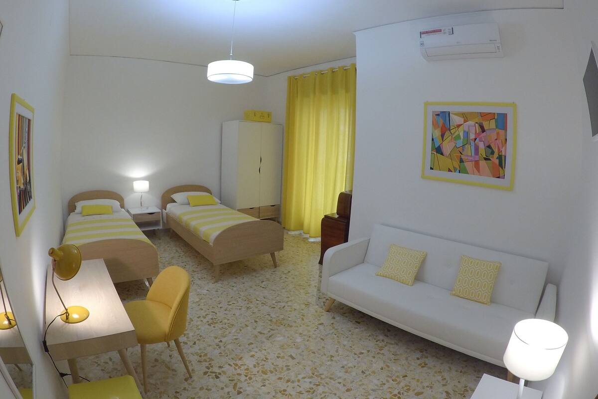 A hotel room with two single beds, a sofa, a desk with a chair, a mirror, and a painting on the wall.