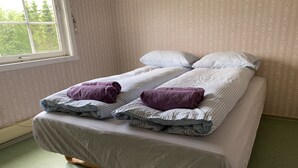 Cama doble con una colcha blanca y dos toallas moradas, sobre un suelo verde y una ventana con vistas a árboles.