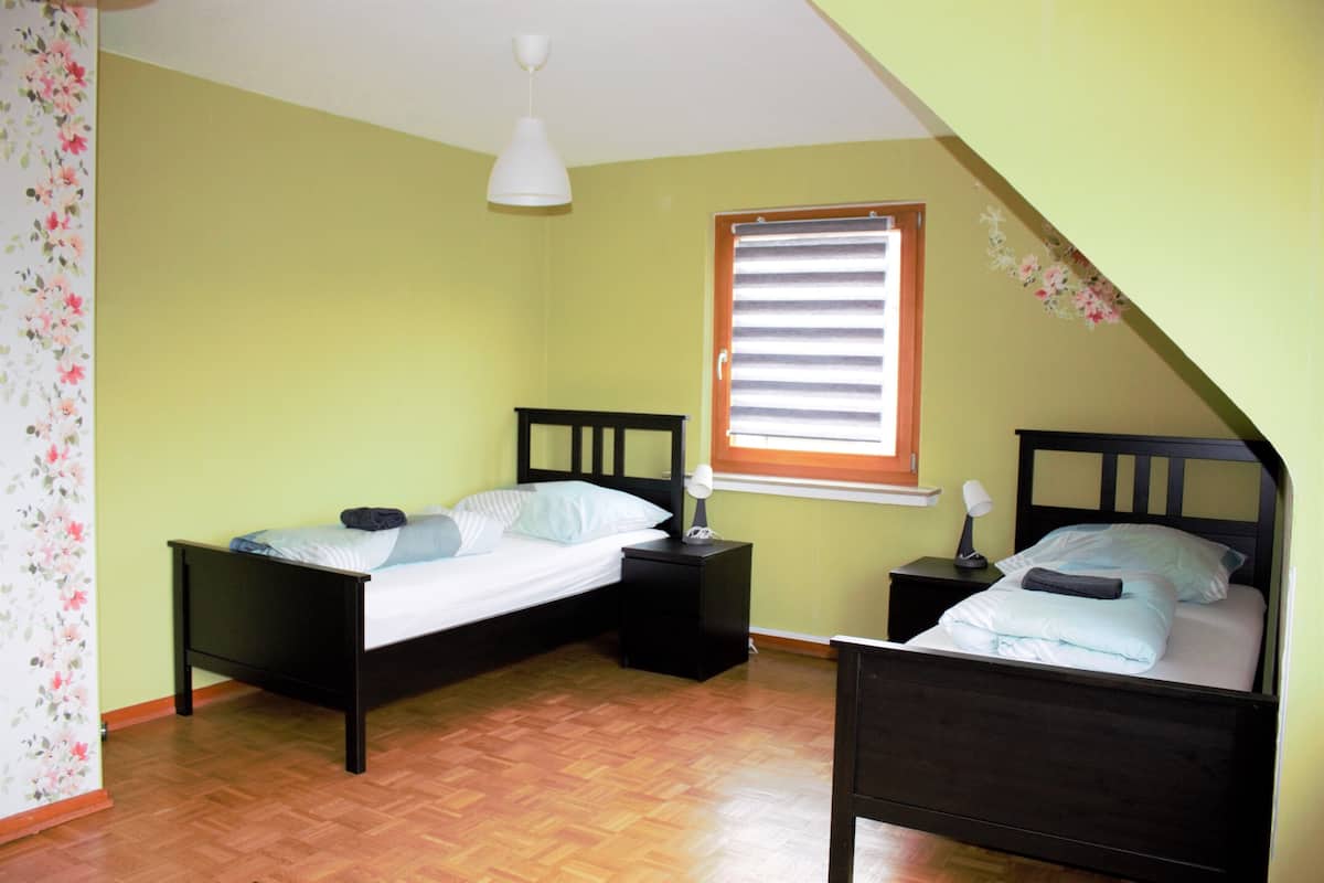 Quarto com duas camas, janela com persianas, piso de madeira e luminária de teto branca.