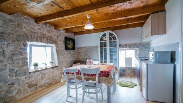 Une cuisine rustique aux murs de pierre, avec des poutres en bois et un coin repas comprenant des chaises blanches et une table.