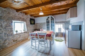 A rustic kitchen with stone walls, wooden beams, and a dining area with white chairs and a table. - Apartment Imota Stone (Proložac)