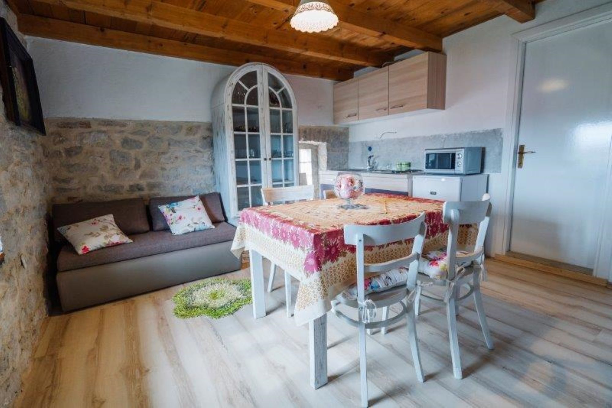 A cozy room with a stone wall, wooden ceiling, a dining table with chairs, a sofa, and a microwave in the kitchen area.