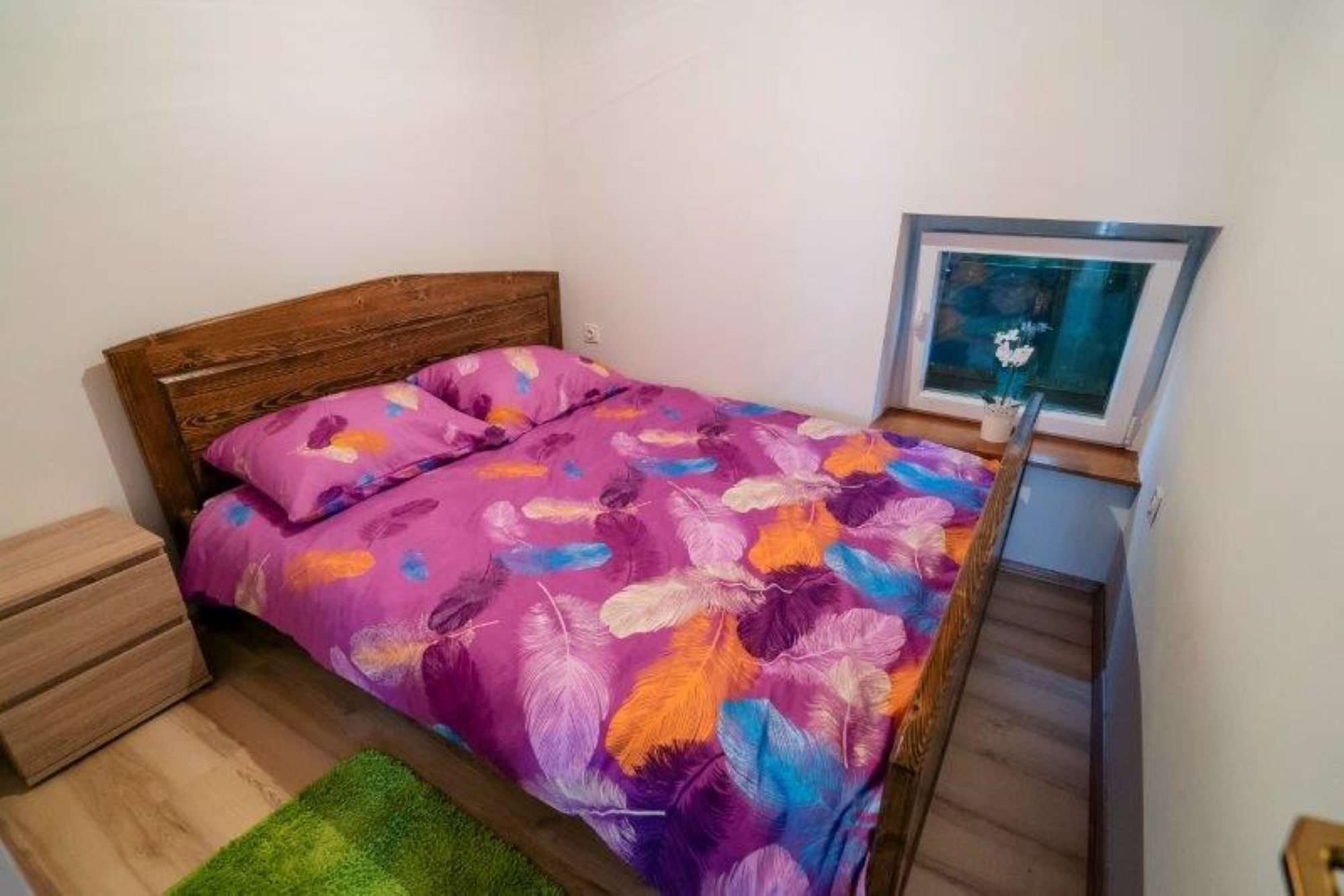 Een slaapkamer met een houten hoofdbord, een paars bloemenbedekking, een groen vloerkleed en een ingelijst beeld aan de muur.