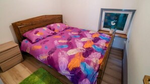 Une chambre avec une tête de lit en bois, une couvre-lit à motifs floraux violets, un tapis vert et un tableau encadré accroché au mur.