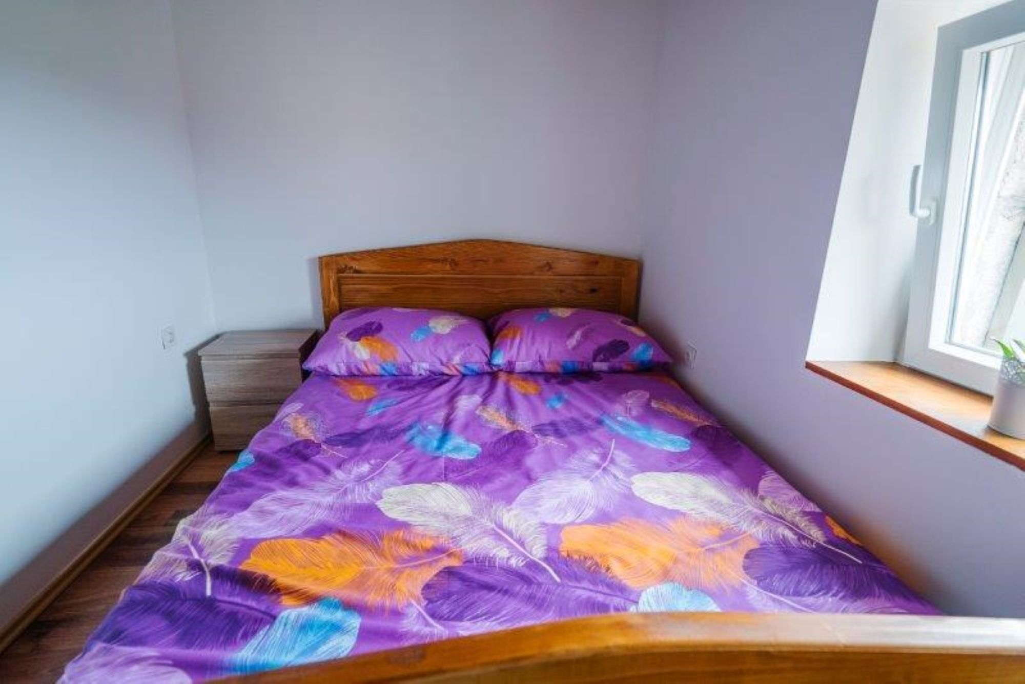 A small bedroom with a wooden headboard, a purple feather-patterned bedspread, and a window with a plant on the sill.