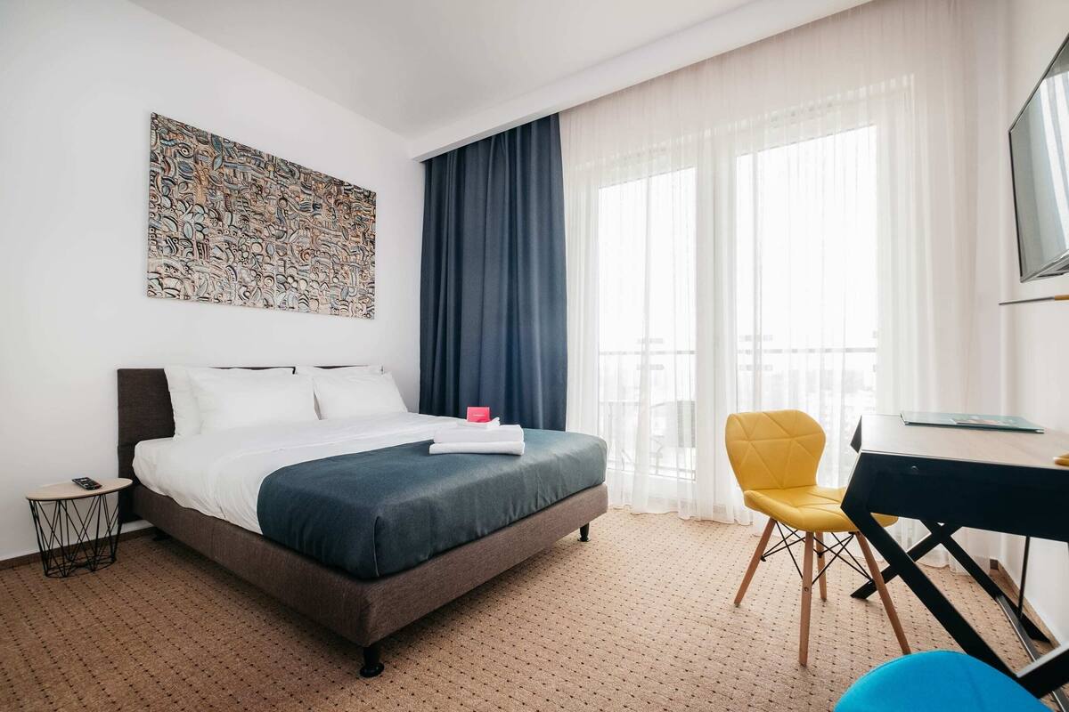 Une chambre d’hôtel avec un lit, un bureau, une chaise, un tableau au mur et une fenêtre avec des rideaux.
