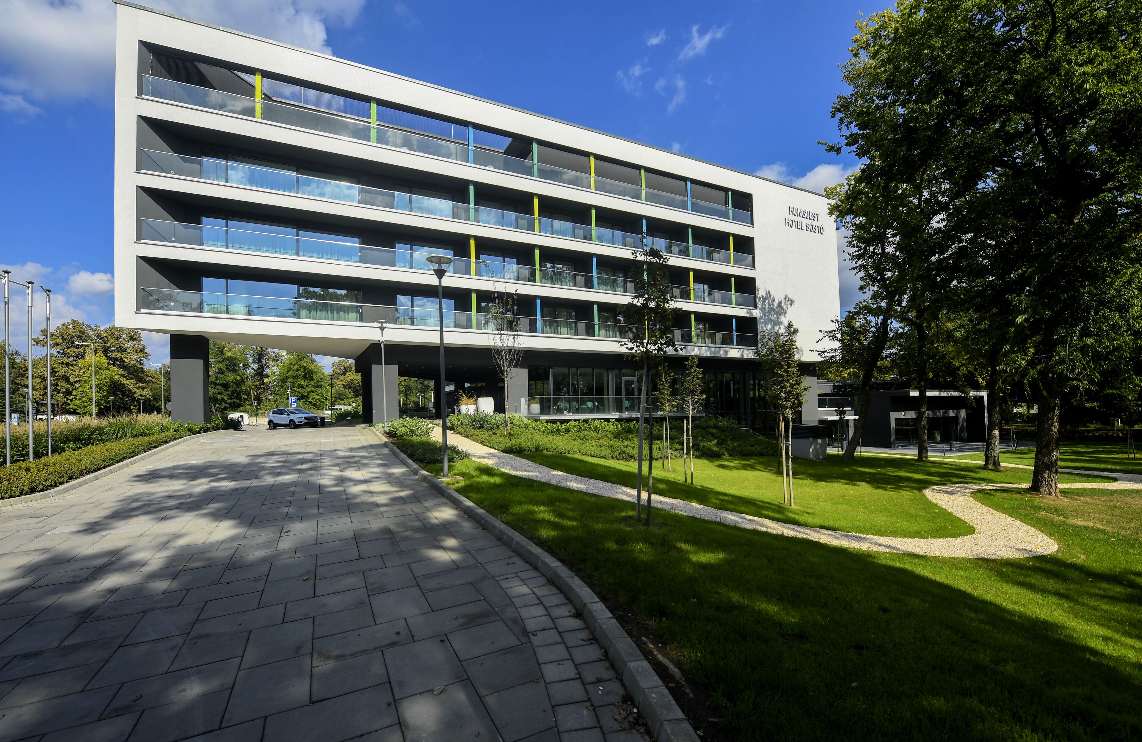 A modern hotel building with multiple floors and balconies, surrounded by well-maintained greenery and a paved walkway.
