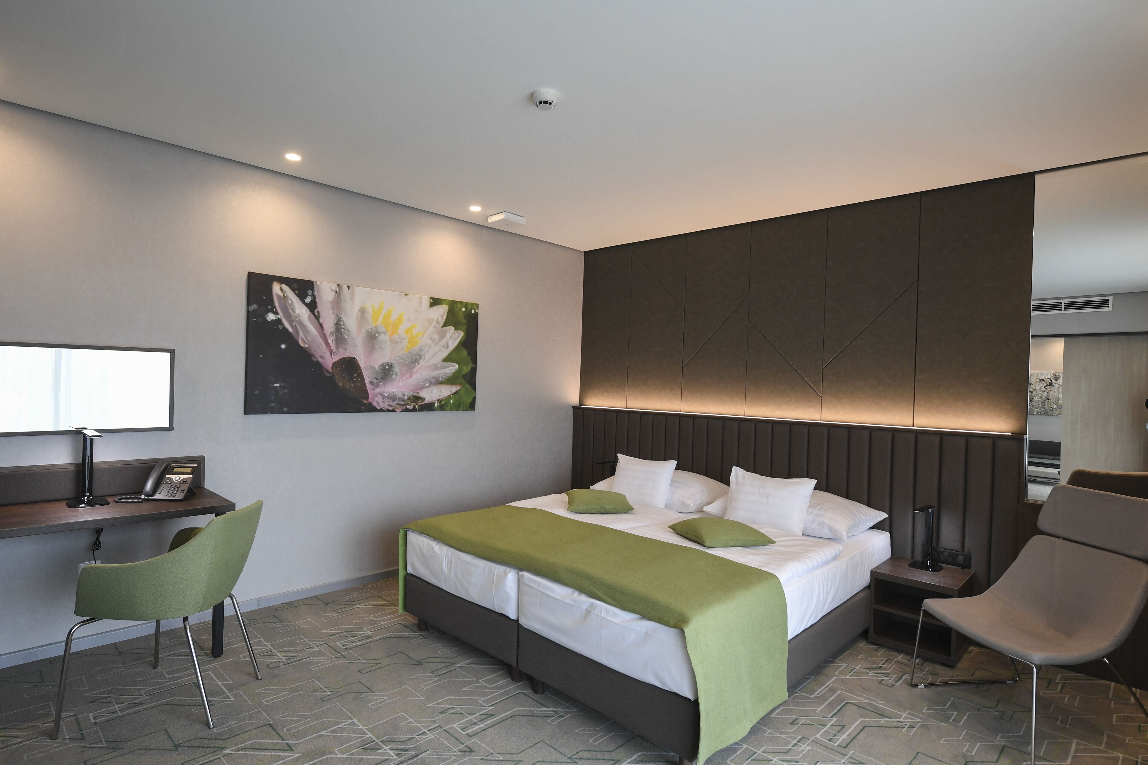 A modern hotel room with a large bed, a desk, two chairs, and a painting on the wall.
