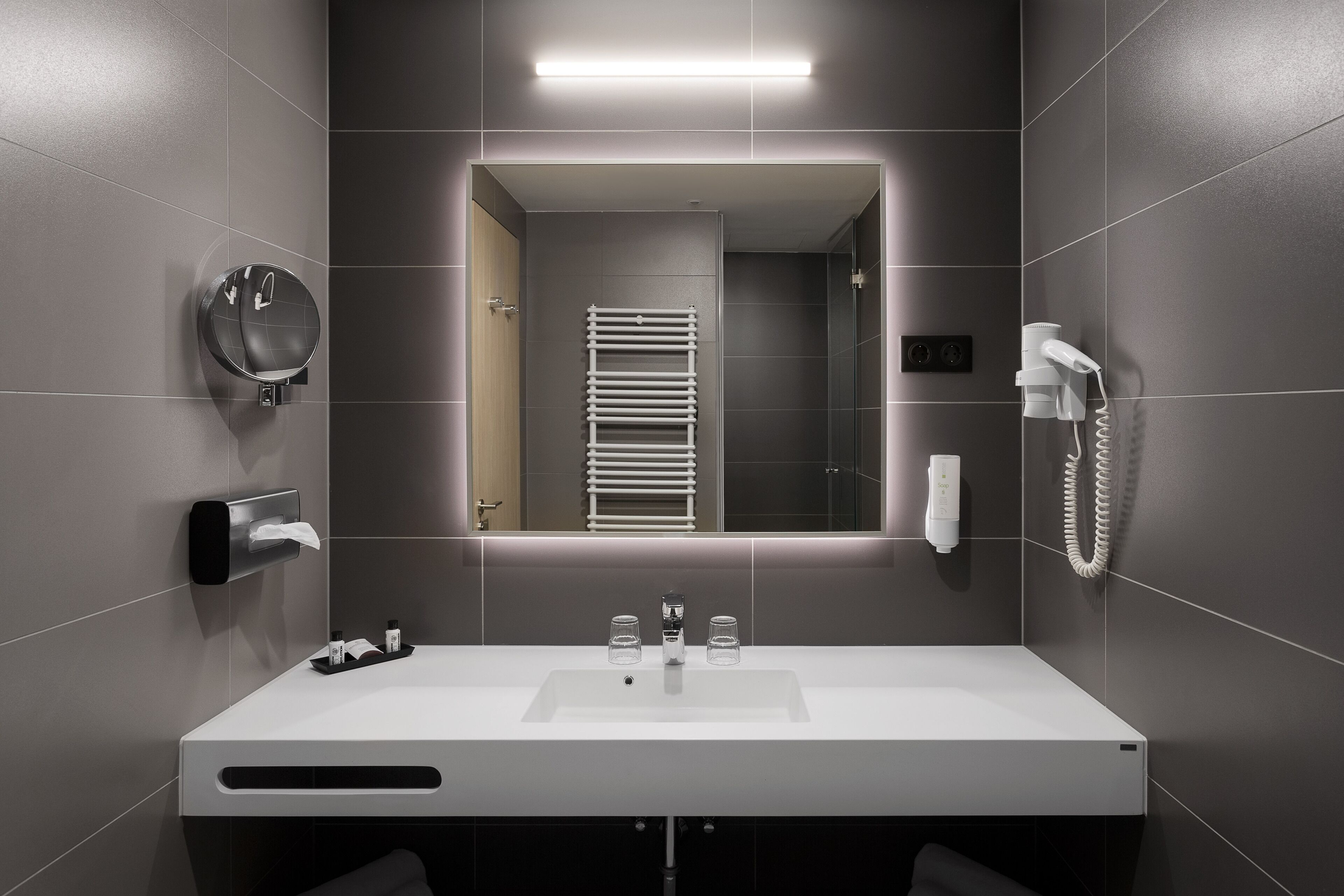 A modern bathroom with a white sink, a towel dispenser, a mirror, and a hairdryer.