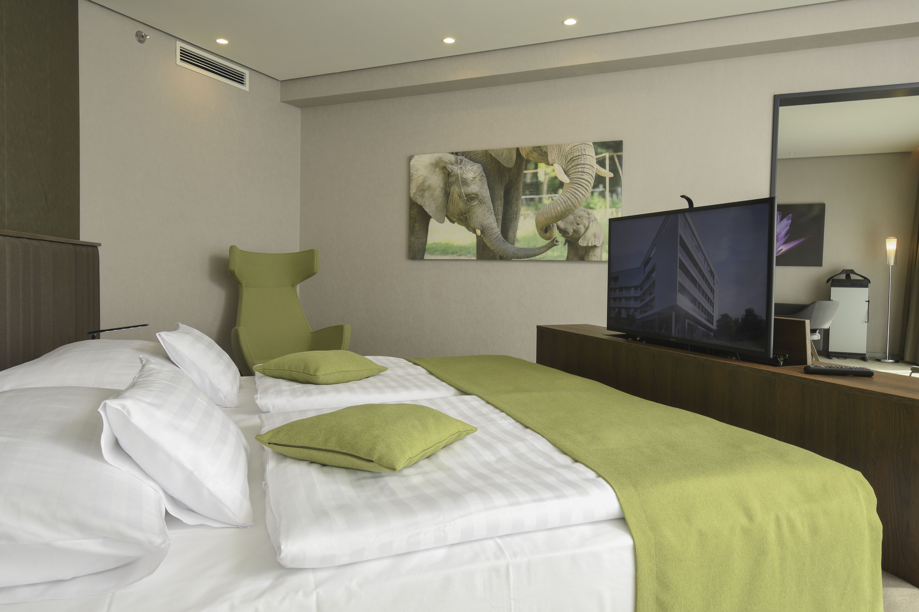 A hotel room with a bed, green pillows, a television on a wooden stand, and a painting of elephants on the wall.