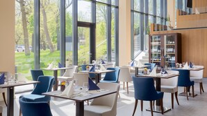 A modern dining area with large windows, blue and white chairs, and neatly arranged tables with menus. - Hunguest Hotel Sóstó (Nyiregyhaza)