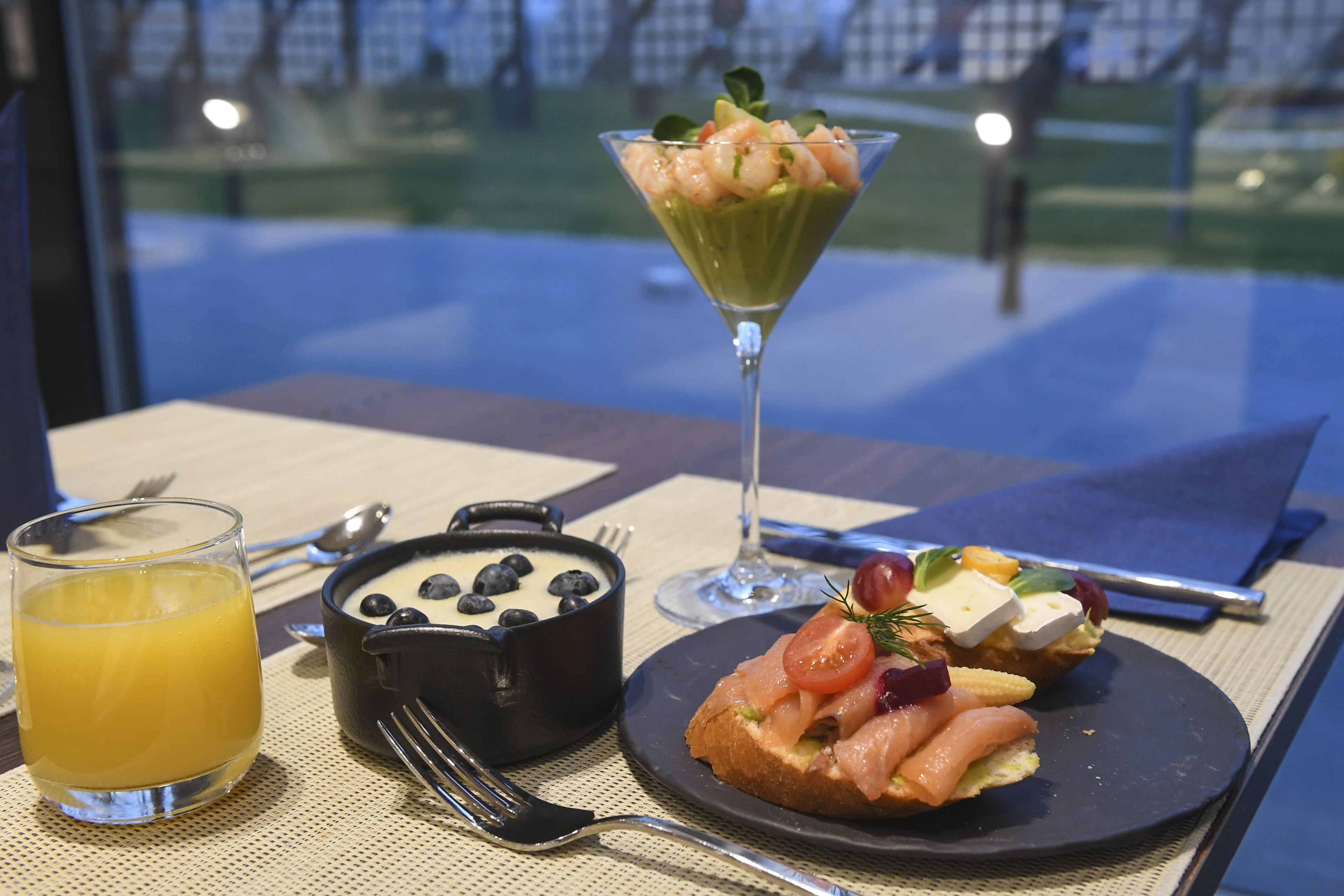 Ein Frühstückstisch mit Orangensaft, einer Schale Blaubeeren, einem Martiniglas mit Garnelen und einem Teller mit Lachs und Gemüse.