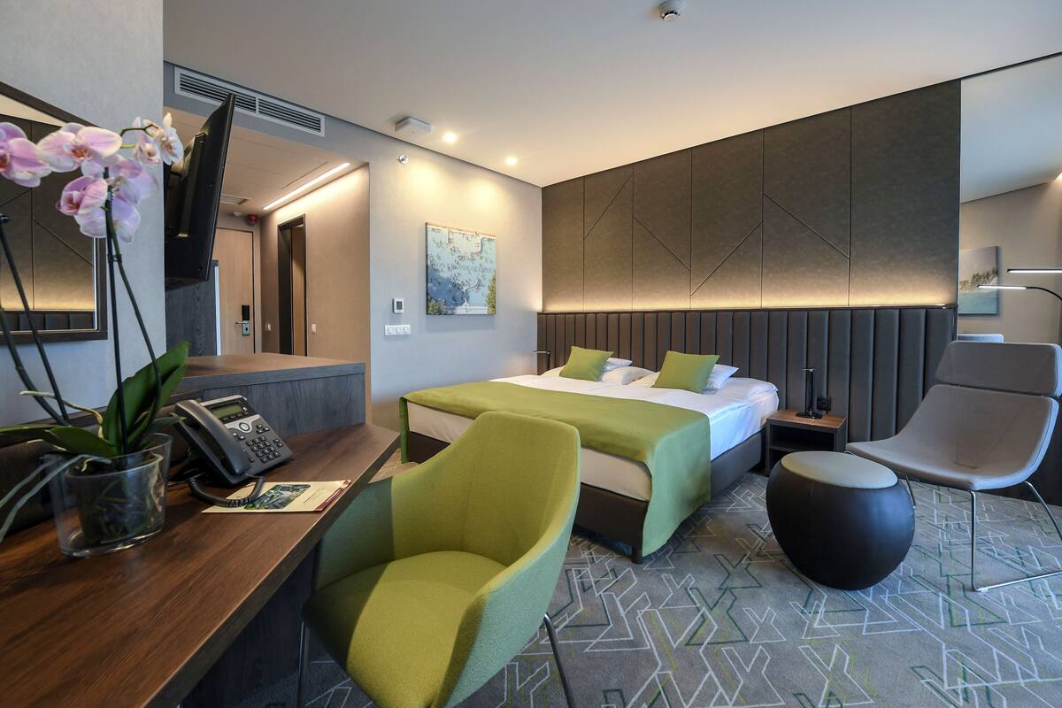 A modern hotel room with a bed, two green chairs, a desk with a phone and a plant, and a map on the wall.