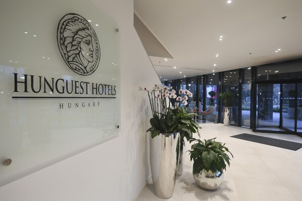 Un moderno vestíbulo de hotel con un gran letrero de HUNGUEST HOTELS HUNGARY, plantas en macetas grandes y una entrada de vidrio.