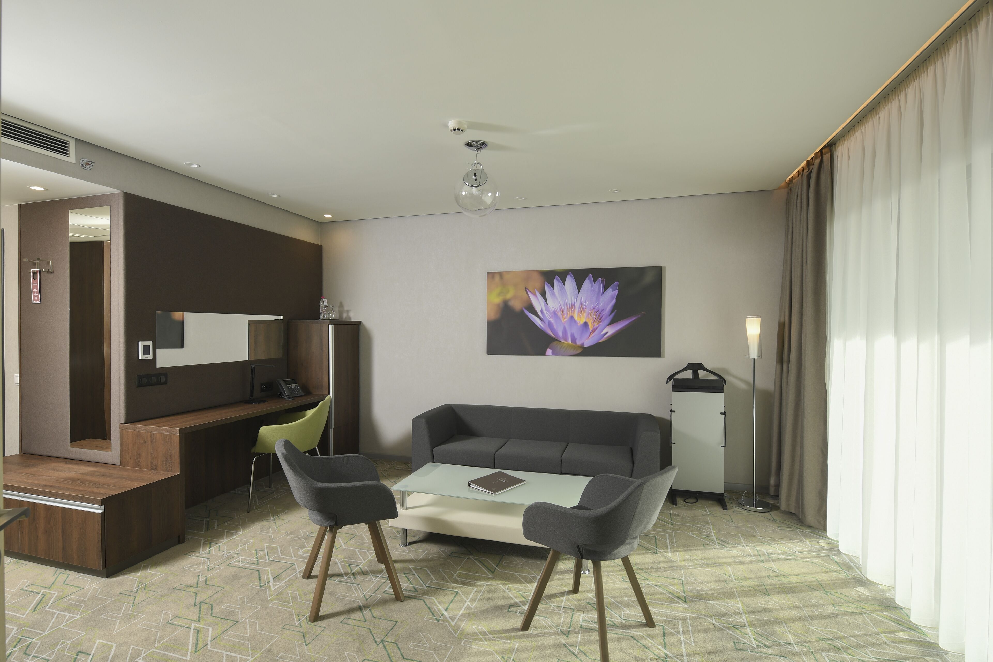 Ein modernes Hotelzimmer mit einer Couch, zwei Sesseln, einem Couchtisch, einem Schreibtisch mit Stuhl, einem großen Fenster mit Vorhängen, einem wandmontierten Fernseher und einem Blumenbild.