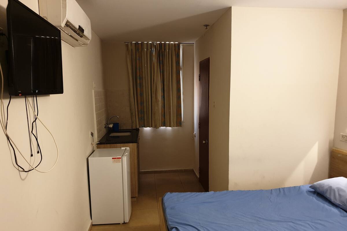 Una habitación pequeña y funcional con cama, kitchenette y televisor montado en la pared.