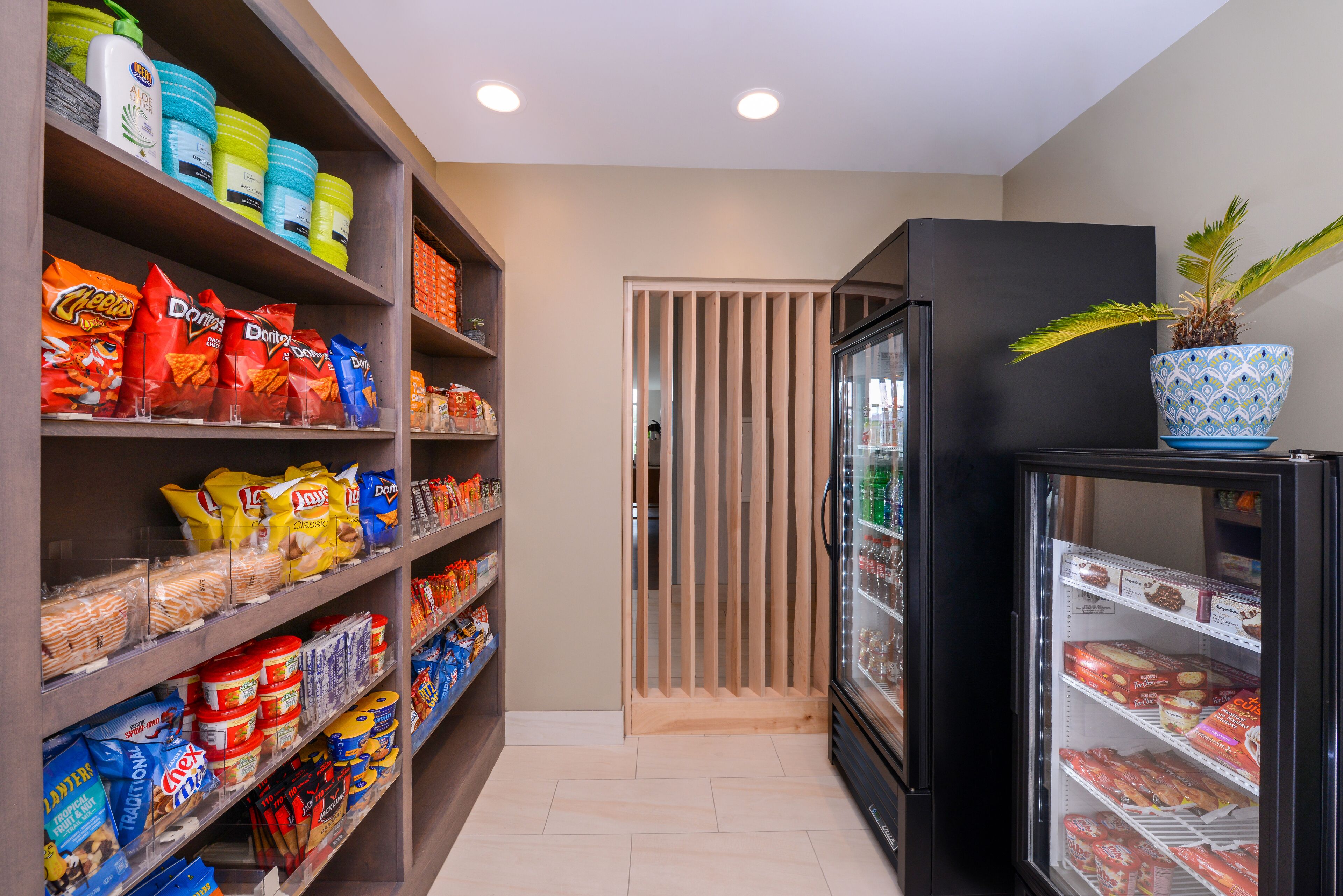 A snack and beverage section with shelves stocked with various snacks and drinks, a refrigerator with a potted plant on top, and a wooden partition in the background.