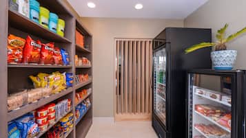 A snack and beverage section with shelves stocked with various snacks and drinks, a refrigerator with a potted plant on top, and a wooden partition in the background.