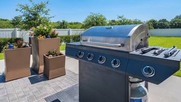 A stainless steel barbecue grill with four burners and a built-in thermometer, positioned on a paved patio with potted plants nearby.