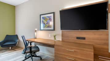 Un espace de bureau moderne avec un bureau en bois, une chaise de bureau noire, une œuvre d’art abstraite encadrée, un téléviseur à écran plat et un vase avec des fleurs blanches.